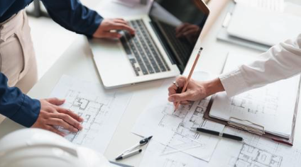 Two people review documents spread across a table. One person types on a laptop while the other writes or draws on the plans with a pencil. A white hard hat is visible in the corner, suggesting a fieldwork meeting is in progress.